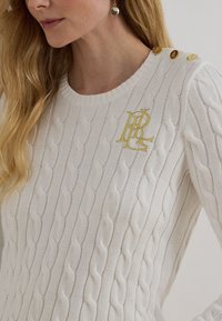 Bela pulover z pletenim vzorcem in z zlatim vezanim logotipom na prsih ter tremi zlatimi gumbi na rami, ki ga nosi oseba z dolgimi blond lasmi.