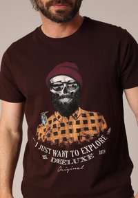 Camiseta marrón con un gráfico de un skull con gafas y barba, vistiendo una camisa a cuadros naranja y un gorro rojo, acompañado de una ilustración de un pájaro.