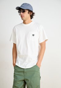White cotton t-shirt with short sleeves and a chest pocket featuring a heart design. Model wears gray cap and sunglasses, paired with green cargo pants.