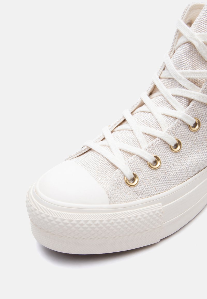 Primo piano di una sneaker bianca alta con tessuto in tela, lacci bianchi infilati in occhielli dorati e punta e suola in gomma.