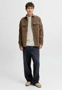 Veste en flanelle marron avec deux poches poitrine, portée sur un pull crème, assortie à un jean sombre ample et des chaussures beiges.