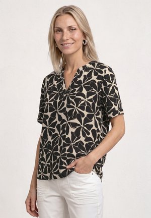 Femme blonde souriante, portant un chemisier à manches courtes à motif floral noir et beige et un pantalon blanc, avec une main dans la poche.