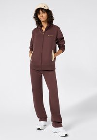 Bruine zip-up jas met zijzakken, gecombineerd met bijpassende broek. Een beige pet completeert de outfit. Stof lijkt zacht en heeft een comfortabele pasvorm.