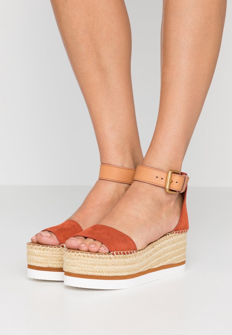 Jambes portant des sandales compensées en daim couleur rouille avec des brides beige à la cheville et des semelles compensées en espadrille tissée sur un sol blanc.