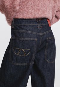 Blugi din denim închis la culoare, cu talie înaltă, având cusături galbene și un design unic în formă de inimă pe buzunarele din spate. Etichetă roșie simplă vizibilă.