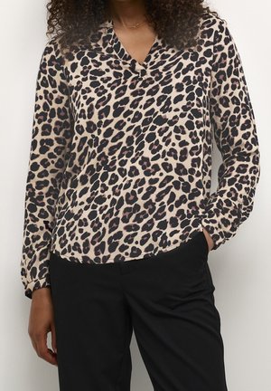 Personne portant un chemisier à manches longues beige et noir à imprimé léopard avec un pantalon noir, debout devant un fond uni clair.