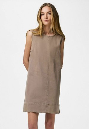 Mladá žena s dlhými blond vlasmi v nosiaca bezrukávové šaty taupe až po kolená, stojaca s rukami za chrbtom na bielom pozadí.