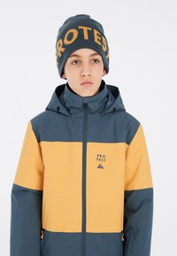 Farbblockierte Jacke in Dunkelblau und Gelb, mit einem Reißverschluss vorne und Kapuze. Graue Mütze mit "PROTEST" in orangefarbener Schrift.