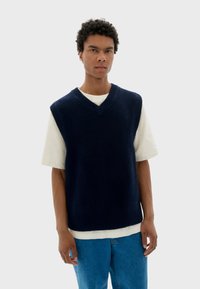Pull sans manches en tricot bleu marine avec un design en V, présentant des détails côtelés et porté par-dessus un t-shirt blanc. Jean en denim visible en dessous.