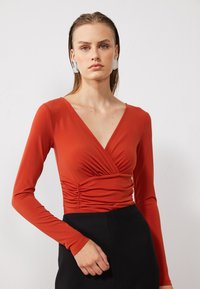 Rust-colored long-sleeve top with a V-neck and gathered detail at the waist, made of smooth, stretchy fabric. Black skirt completes the outfit.