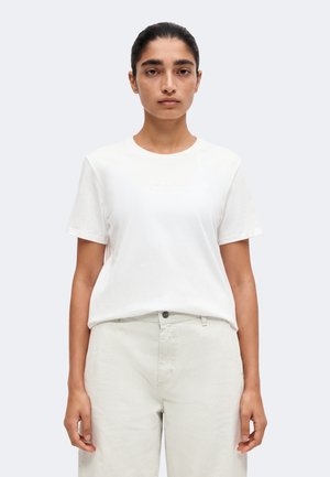 Femme aux cheveux foncés attachés en arrière, portant un t-shirt blanc uni à manches courtes et un pantalon clair rayé, debout devant un fond clair uni.