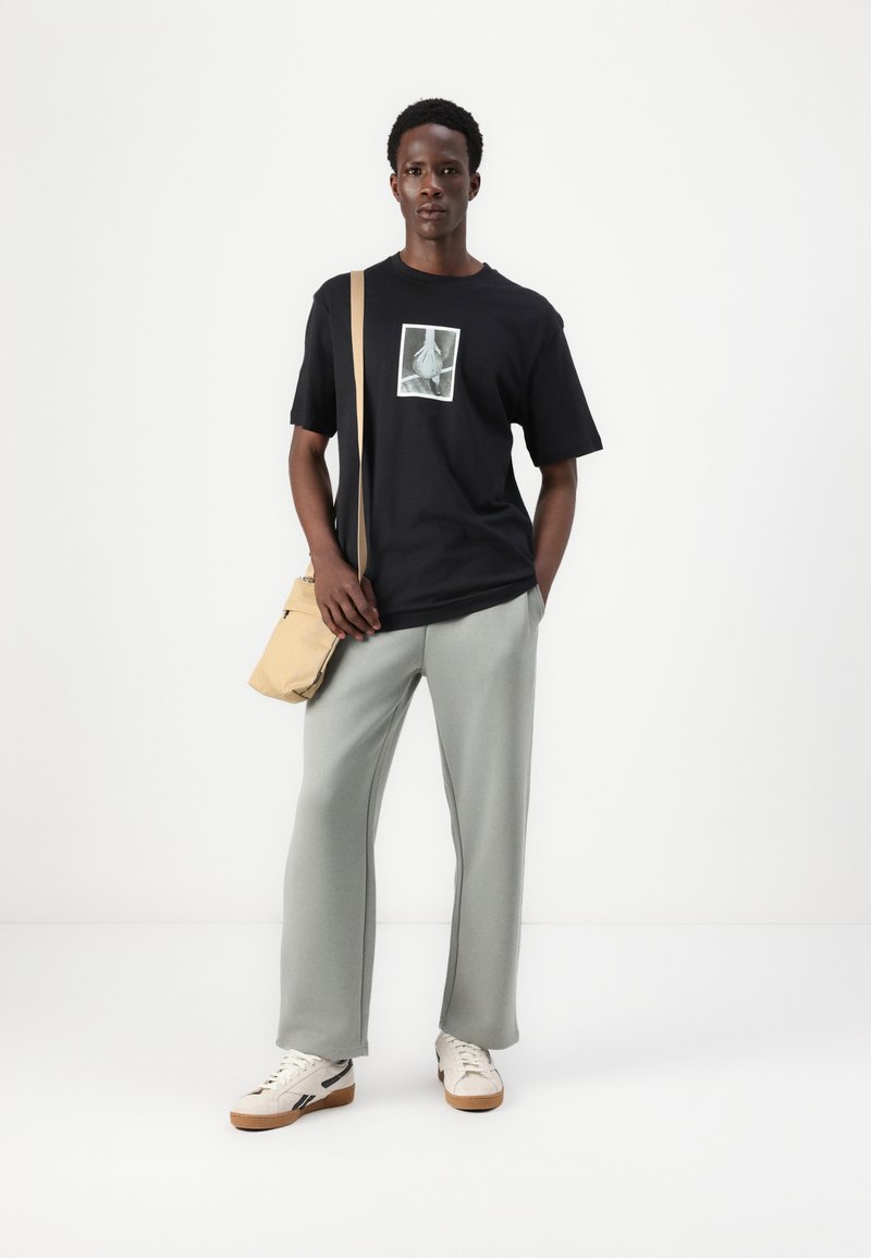 Sort T-shirt med grafisk tryk, lysegrå sweatpants, creme sneakers og en beige crossbody taske. Simpelt design med afslappet pasform.