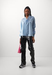 Lyseblå bluse med knytning ved halsen, lange ærmer, parret med blanke sorte bukser og en rosa håndtaske, med sporty sneakers.