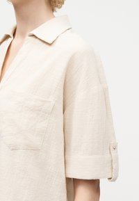 Γυναίκα που φοράει μια χαλαρή, ανοιχτή μπεζ υφασμάτινη μπλούζα με γιακά, τσέπη στο στήθος και κοντά μανίκια γυρισμένα προς τα πάνω, πάνω σε απλό φόντο.