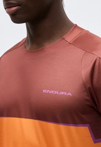Κοντινό πλάνο ενός ατόμου που φοράει αθλητική μπλούζα σε καφέ και πορτοκαλί, με το λογότυπο "ENDURA" σε ανοιχτό μωβ στο στήθος.