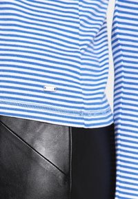 Gestreiftes blaues und weißes Oberteil mit kurzen Ärmeln, das ein kleines Logo-Label und einen gerippten Saum aufweist, kombiniert mit schwarzem strukturiertem Stoff darunter.