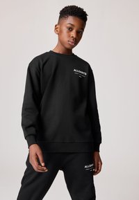 Teenager s krátkými kudrnatými vlasy ve černé mikině AllSaints a černých kalhotách na jednoduchém světlém pozadí.