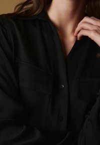 Černá košile na knoflíky s límečkem, dvěma náprsními kapsami a texturovanou tkaninou. Má volný střih a dlouhé rukávy.