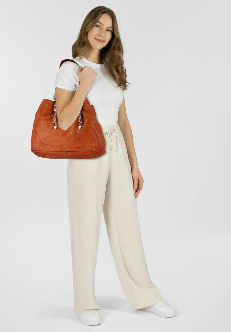 Sac à main en cuir orange avec des accents de poignées en perles, présentant un design décontracté et une grande ouverture, tenu par une personne portant un pantalon clair.