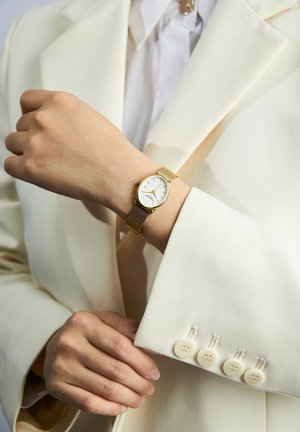 Zlatni sat s mrežastim remenom i bijelim ovalnim brojčanikom, s minimalističkim oznakama sati. Nošen na ruci odjevenoj u slonovačku sako.