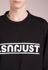 Sudadera negra con cuello redondo, que presenta un gran gráfico de texto blanco que dice "JUST." El material parece suave con una textura sutil.