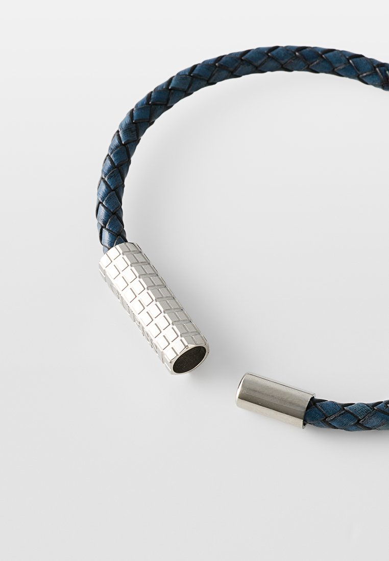 Bracelet en cuir bleu tressé avec un fermoir en argent texturé et des détails métalliques, présentant un motif géométrique cylindrique.