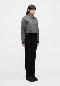 Žena stojaca, oblečená v krátkej sivej bunde na zips, čiernom tričku s dlhým rukávom, čiernych nohaviciach s rovnými nohavicami a čiernych masívnych topánkach na bielom pozadí.