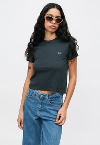 Svart avkortet t-skjorte med hvit "obey" logo, kombinert med høy midje blå denimjeans. Modell iført grønne katteøyne solbriller.