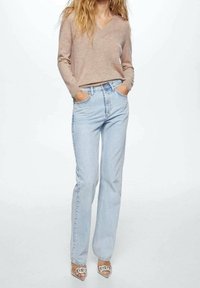 Hellblaue, hoch taillierte Jeans mit geradem Bein, kombiniert mit einem beigen V-Ausschnitt-Pullover und transparenten Sandalen mit Absatz.