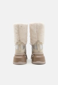Bottes en fourrure beige avec un tige slim, en suède texturé, accents en maille métallique et une semelle marron superposée. Talon renforcé avec une languette.