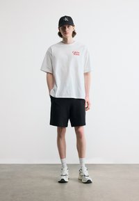 Νέος άνδρας στέκεται φορώντας λευκό t-shirt Calvin Klein, μαύρο σορτς, μαύρο καπέλο, λευκές κάλτσες και λευκά αθλητικά παπούτσια σε γκρι πάτωμα και λευκό φόντο.