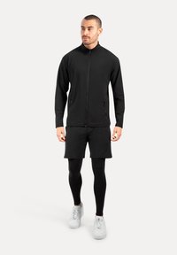 Hombre vestido con chaqueta negra con cremallera, pantalones cortos negros, mallas negras y zapatillas blancas, de pie contra un fondo blanco.