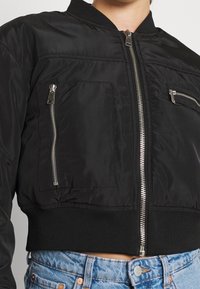 Black bomber jacket with a cropped design, featuring two zippered pockets, a ribbed waistband, and smooth fabric texture. Silver hardware accents.