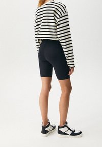 Personne portant un short de cycliste noir, des baskets montantes noir et blanc, et une chemise blanche à manches longues avec des rayures horizontales noires, debout sur un sol blanc.