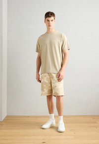 Beige cotton t-shirt with short sleeves, paired with beige camo-pattern shorts. Model wears white sneakers and crew socks, standing on wooden floor.