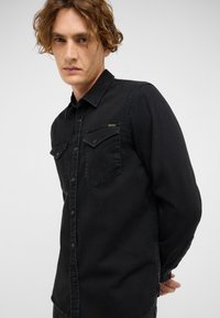 Juodas džinsinis marškinis su mygtukų užsegimu, dviem priekinėm kišenėm ir klasikinio tipo apykakle. Pabrėžia subtilų siuvinėjimą ir modernų siaurą fasoną.