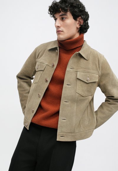 Ταραντένιο σουέτ μπουφάν με κλείσιμο κουμπιού και δύο τσέπες στο στήθος, φορεμένο πάνω από ριμπ τζέρσεϊ σε χρώμα σκουριάς. Μαύρα παντελόνια ολοκληρώνουν το σύνολο.