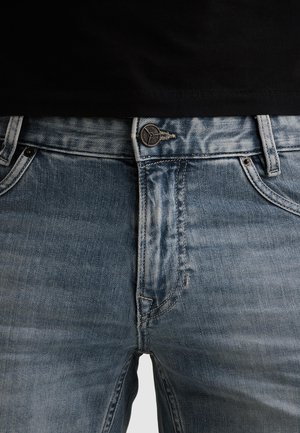 Nahaufnahme des Taillenbereichs einer hellblauen Jeans aus Denim mit einem Metallknopf und Gürtelschlaufen, getragen mit einem schwarzen Hemd.