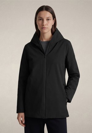 Mujer con cabello castaño liso que lleva un abrigo negro con cremallera y cuello alto, con las manos parcialmente en los bolsillos, de pie contra un fondo liso.