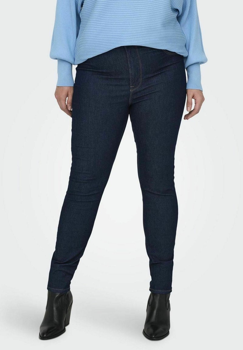 Blugi skinny de culoare albastru închis, din denim, cu un elastic standard și detalii de cusătură subtile, asortați cu cizme negre până la glezne.