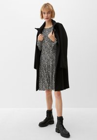 Schwarzer Mantel mit hohem Kragen über einem floral gemusterten Kleid in Schwarz und Weiß. Ein Paar schwarze, geschnürte Kampfkuschen mit dicken Sohlen.