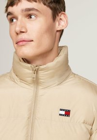 Beige puffer jacket with high collar and zip closure. Features a small rectangular logo patch with navy, white, and red accents on the chest.