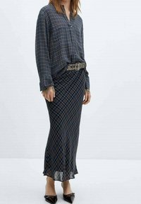 Longue jupe à carreaux bleu foncé avec des accents rouges et verts, assortie à une chemise à carreaux boutonnée. Comprend une ceinture noire cloutée et des talons.