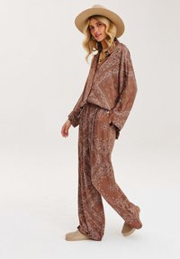 Dvojdielny outfit s hnedým paisley vzorom, ktorý obsahuje voľnú košeľu na gombíky a široké nohavice, doplnený béžovými nazúvacími topánkami a klobúkom.