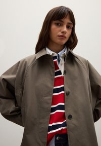 Young woman with long brown hair wears a gray oversized jacket over a red, white, and blue striped shirt and a blue-striped button-up shirt.