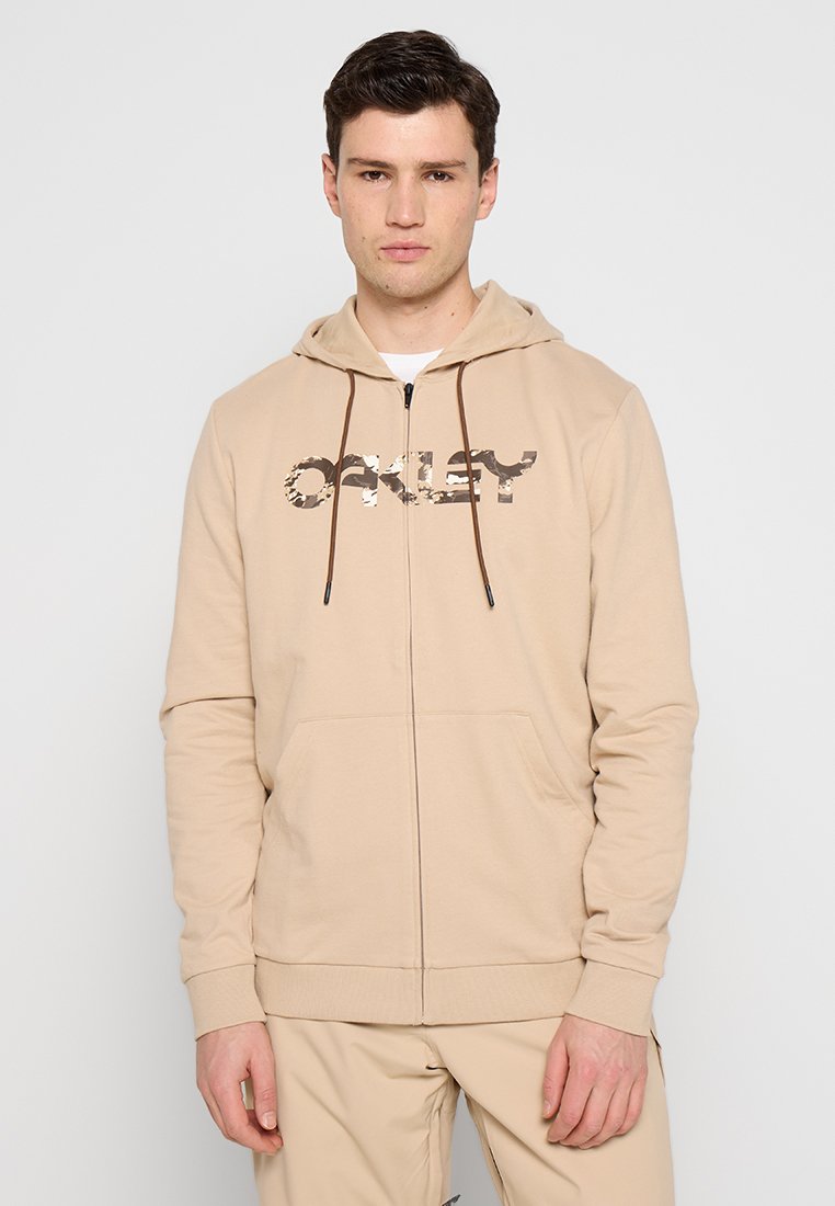 Un jeune homme portant un sweat à capuche zippé beige avec un logo Oakley camo marron et un pantalon beige assorti, sur un fond uni.