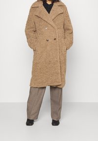 Persona che indossa un cappotto lungo doppiopetto color sabbia con texture, con le mani nelle tasche, abbinato a pantaloni grigi a gamba larga e scarpe nere.