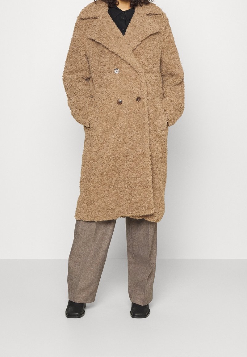 Persona che indossa un cappotto lungo doppiopetto color sabbia con texture, con le mani nelle tasche, abbinato a pantaloni grigi a gamba larga e scarpe nere.