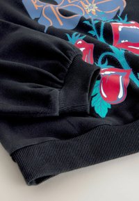 Sort sweatshirt med ribbede manchetter og søm. Har et farverigt grafisk design med røde roser og blå blade, med detaljeret dekoration.