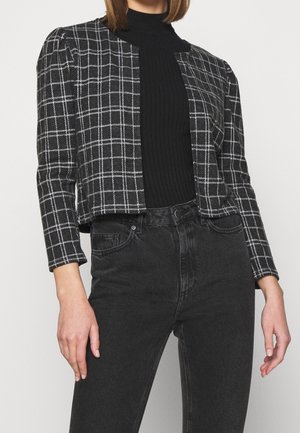 Frau trägt eine schwarz-weiß karierte kurz geschnittene Jacke über einem schwarzen, gerippten Rollkragenpullover mit hoch taillierten schwarzen Jeans.